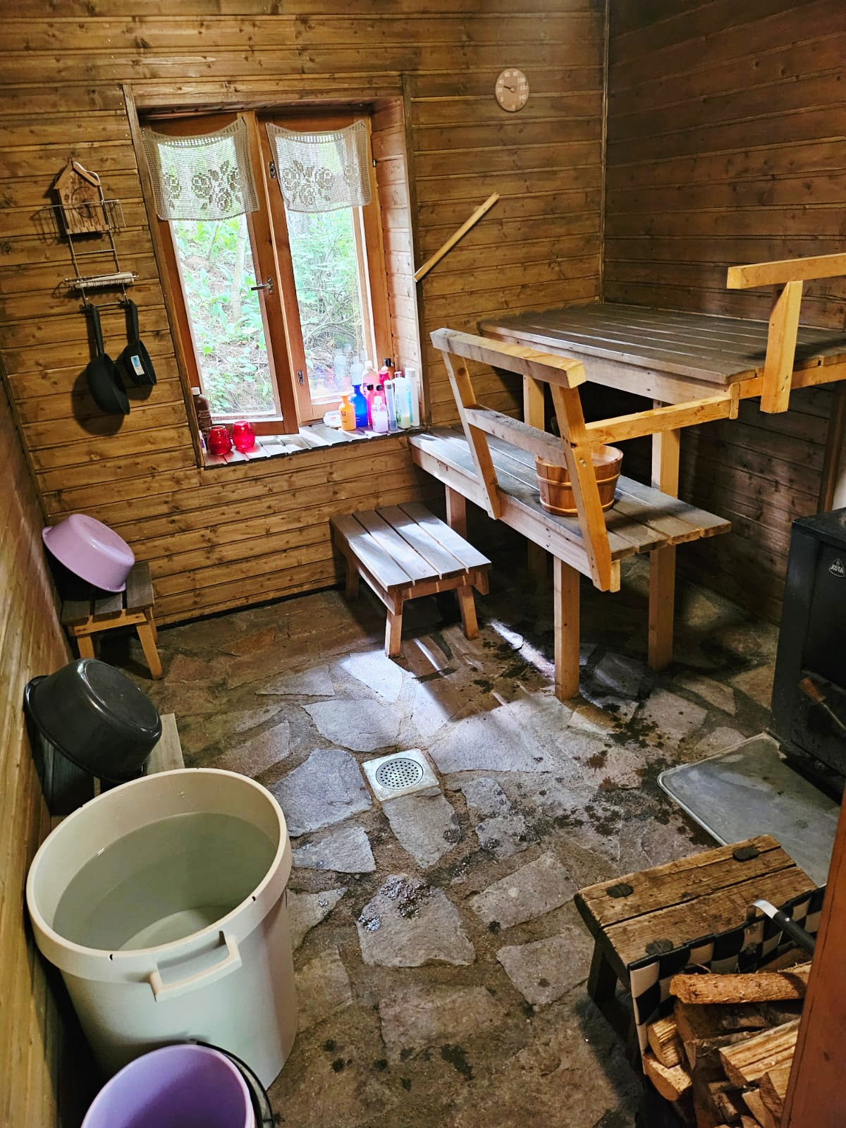 Puinen sauna, jossa penkit, pesuallas, ämpäri ja ikkuna, seinillä pyyhkeet sekä valaistus katossa.
